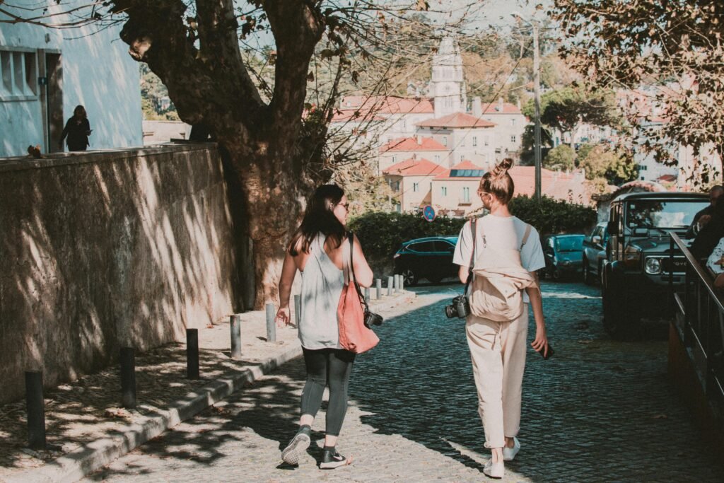 Two women stroll down a picturesque cobblestone street in Portugal, capturing the beauty with their cameras.